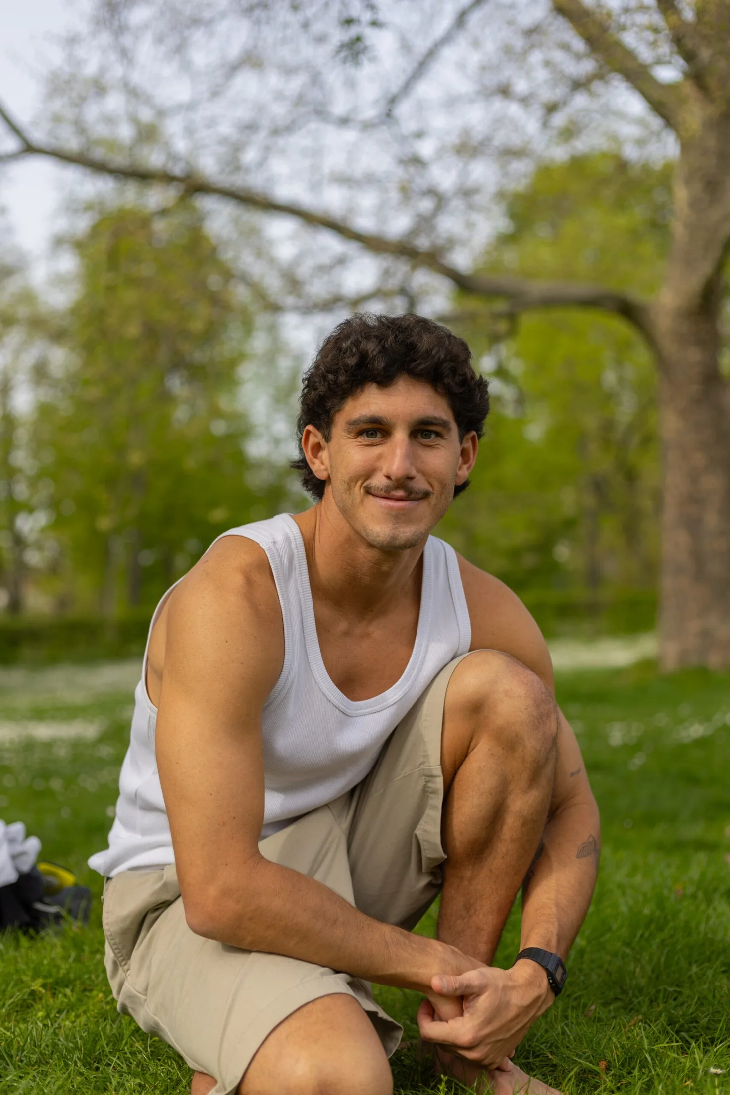 Mann im weißen Tanktop hockt lächelnd im Gras, entspannte Pose im Park
