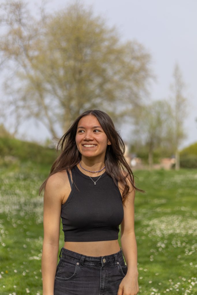 Junge Person mit offenem Haar und schwarzem Crop-Top steht auf einer grünen Wiese voller kleiner Blumen und blickt lächelnd zur Seite.